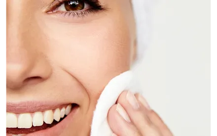 Femme dans sa salle de bain, serviette sur la tête, utilisant un coton pour rincer le gommage double exfoliant utilisé précédemment dans sa routine.