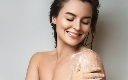 Une femme sous la douche se lave et fait mousser le gel douche sur son corps. De la mousse apparaît entre ses doigts et sur la peau grâce à cette base lavante très douce, pour une peau nettoyée, nette et revitalisée.