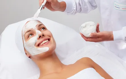 En plein soin en institut, une femme sourit, un masque-crème appliqué sur la moitié du visage. Ce soin en poudre se réhydrate à l'eau à l'aide d'un shaker pour offrir une texture onctueuse. Découvrez le Shaker-Mask Creamy, la solution apaisante spéciale rougeurs.