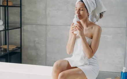 Une femme assise dans sa salle de bain sent un flacon cosmétique. Les effluves de lavande de cette lotion post-épilatoire aux propriétés apaisantes invitent à appliquer ce soin sur les zones épilées