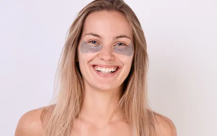Une femme blonde qui sourit avec des patchs « boosters de regard » sous les yeux. La spécificité de ce patch, c'est qu'il est infusé de fleur de pois papillon antioxydant, ce qui lui offre une jolie couleur bleuté avec des incrustations de petites particules de fleur visibles à l'œil.