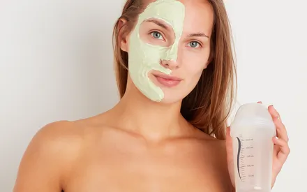 Une femme avec un masque-crème appliqué sur la moitié du visage et un shaker en plastique dans l'autre main, pour faire comprendre le fonctionnement de ce masque en poudre à réhydrater avec de l'eau à l'aide d'un shaker, afin d'obtenir une texture onctueuse. Shaker-Mask Creamy matifiant.