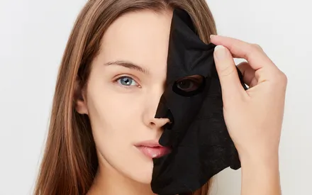 Une jeune femme européenne au sourire. Elle est en train de retirer délicatement un masque en tissu noir au charbon puri-détox de son visage, révélant une peau fraîche, propre et hydratée. 